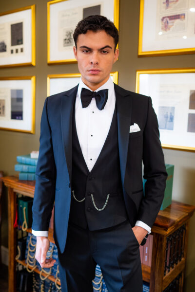 Slim fit classic black dinner suit with traditional black tuxedo jacket, classic black dinner suit trousers and black low cut v shape waistcoat. Features a slim fit 1 button velvet dinner jacket with wide black satin peak lapel. Worn with white studded dress shirt with French cuff shirt and large tom ford style bow tie and white pocket square.