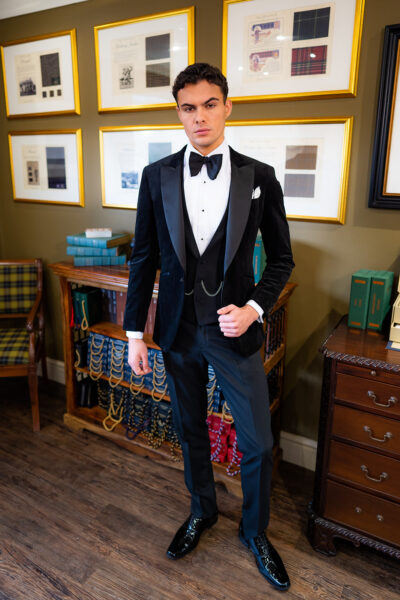 Slim fit classic black velvet dinner suit with traditional black velvet tux jacket, classic black dinner suit trousers and black low cut v shape waistcoat. Features a slim fit 1 button velvet dinner jacket with wide black satin peak lapel. Worn with white studded dress shirt with French cuff shirt and large tom ford style bow tie and white pocket square.